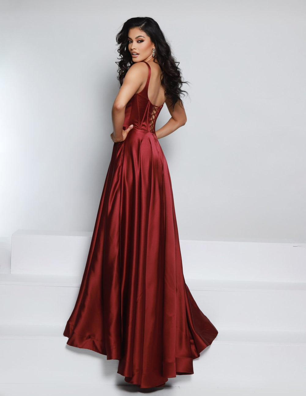 A woman with long, dark, wavy hair poses with her back turned, wearing the 2Cute 25717 floor-length deep red satin gown by 2Cute, featuring a sleeveless fitted bodice and flowing skirt, against a plain white background.