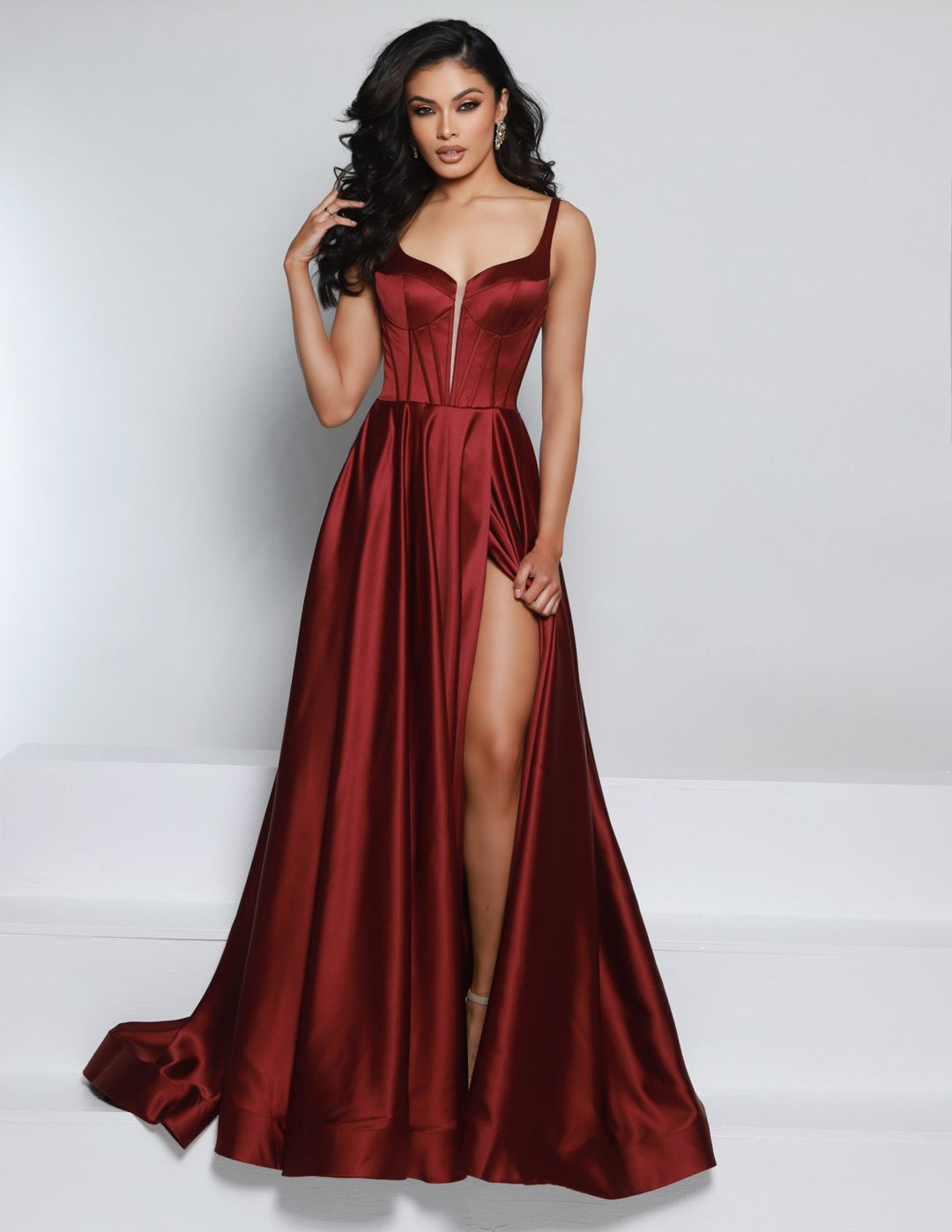 A woman wearing the 2Cute 25717 by 2Cute—a deep red satin evening gown with a fitted bodice, spaghetti straps, and a thigh-high slit—poses against a plain white background. She has long wavy dark hair and high heels.