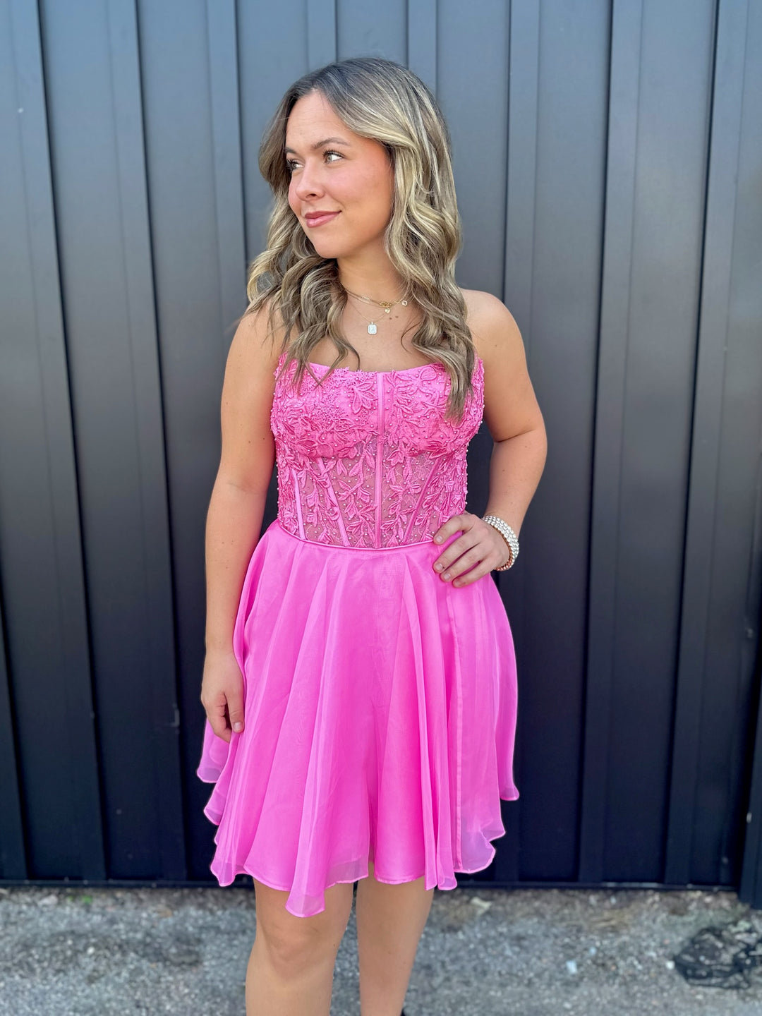 A woman with wavy blonde hair models the Sherri Hill 56502, a strapless lace and chiffon dress in bright pink by Sherri Hill, posing with one hand on her hip before a dark vertical-panel wall.