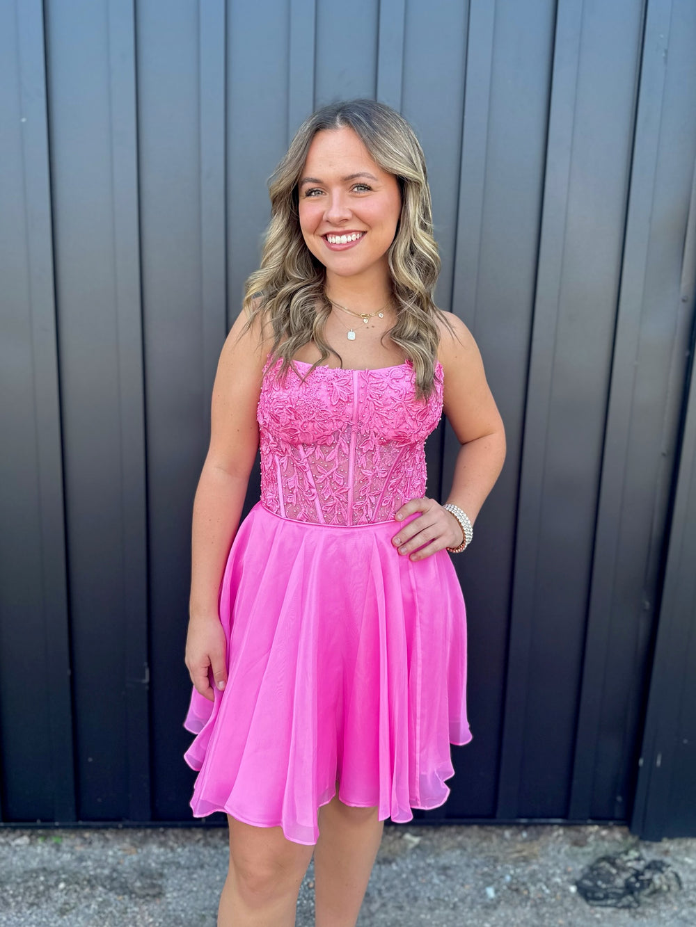 A young woman with wavy blonde hair smiles while wearing the Sherri Hill 56502, a bright pink, sleeveless lace corset dress by Sherri Hill. She stands in front of a dark vertical panel background.