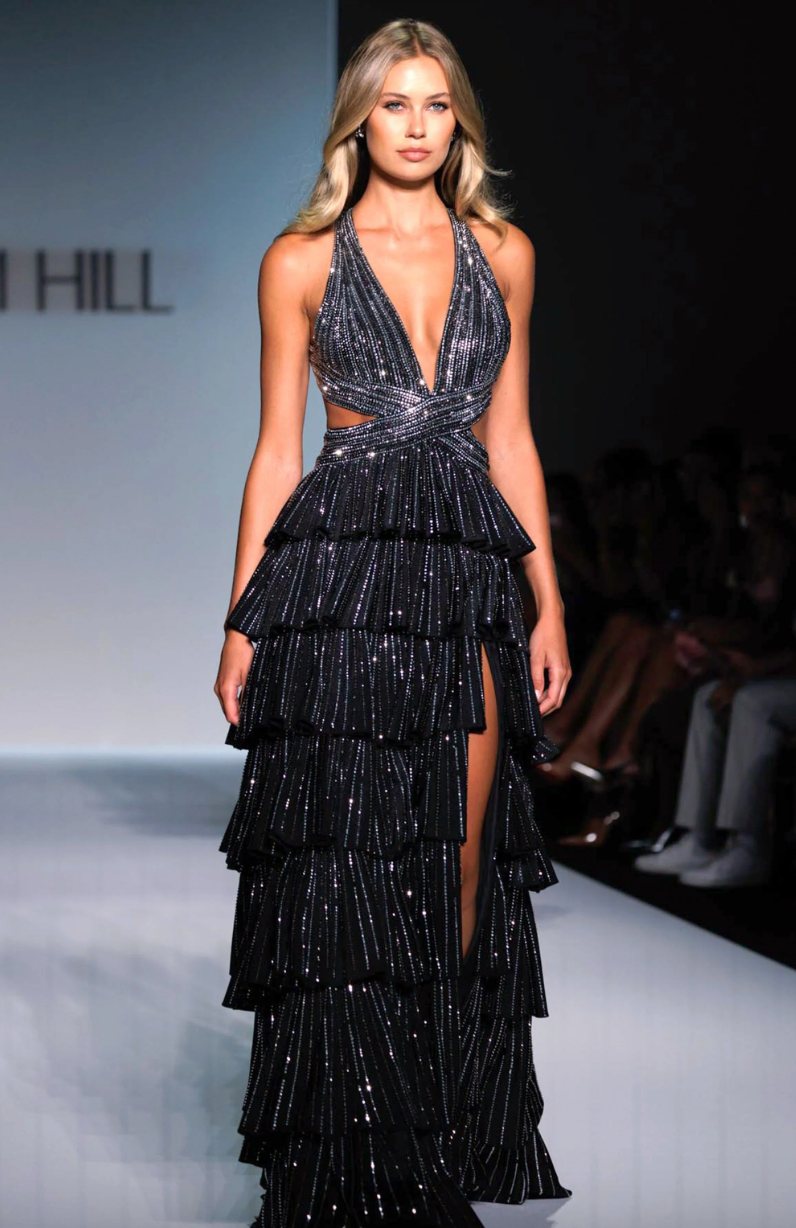 A model walks the runway in the Sherri Hill 57055, a sleeveless black A-line gown with a deep V-neck, sparkling accents, and a dramatic ruffle skirt by Sherri Hill, set against a softly lit fashion show background.