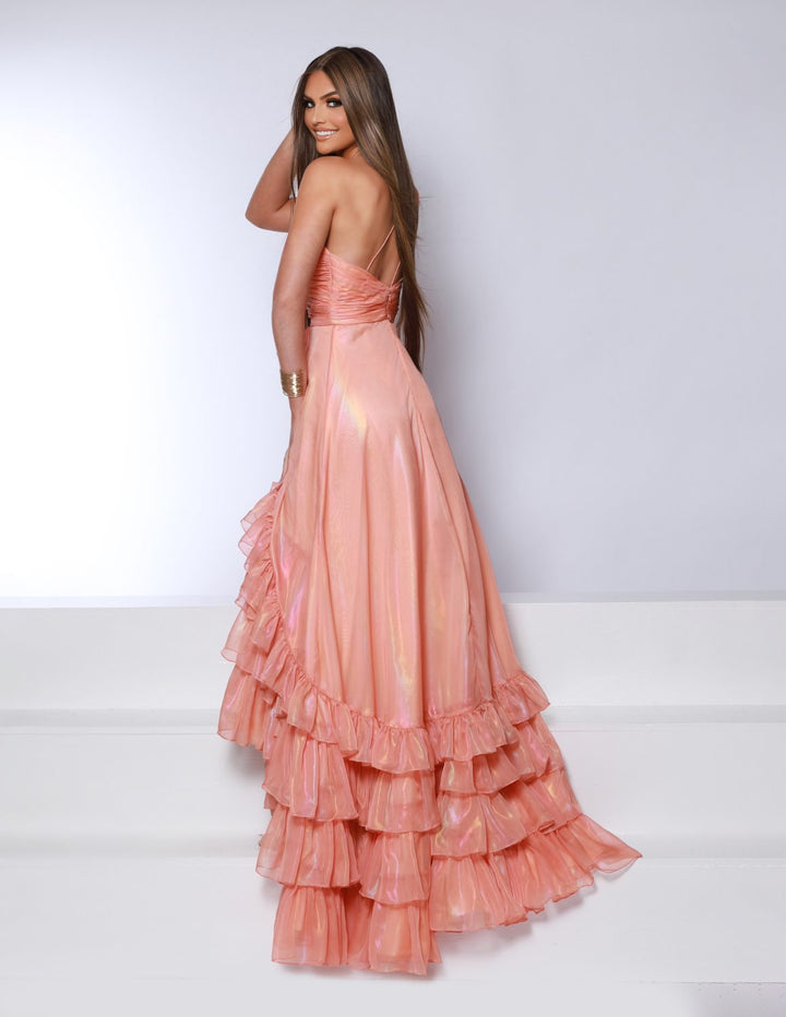A woman with long brown hair in the 2Cute 25350 peach ruffled floor-length gown by 2Cute poses on white steps, looking over her shoulder and smiling. The dress has a low back and layered ruffles at the hem.