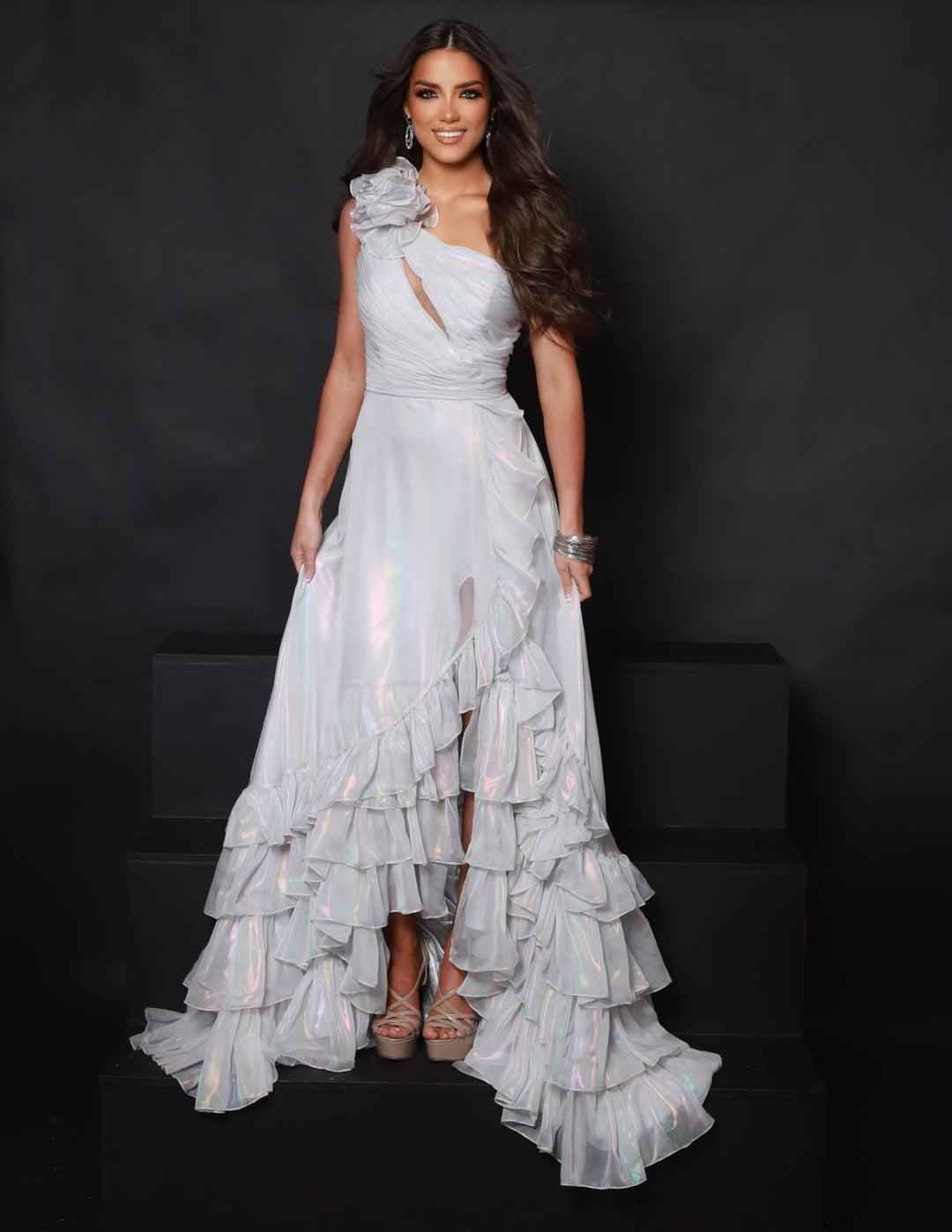 A woman with long brown hair smiles at the camera, wearing the 2Cute 25350 shimmering white ruffled one-shoulder gown and high heels, standing against a plain black background.