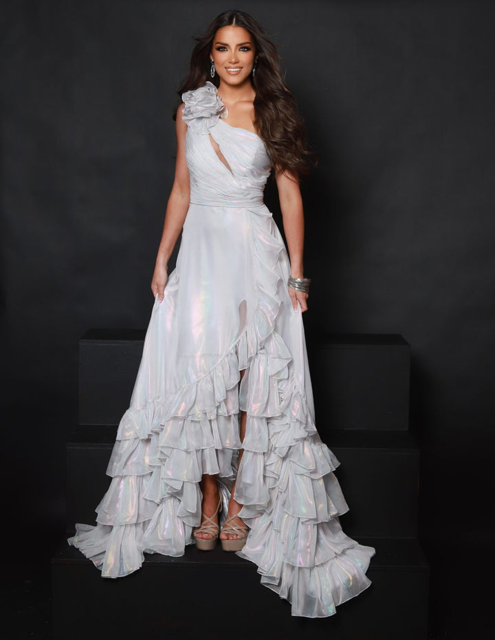 A woman with long brown hair smiles at the camera, wearing the 2Cute 25350 shimmering white ruffled one-shoulder gown and high heels, standing against a plain black background.
