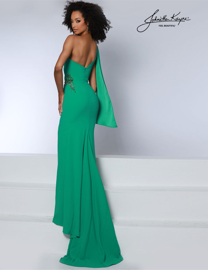 A woman with curly hair poses against a white background in the Johnathan Kayne 2864, a vibrant green, floor-length one-shoulder gown with a long drape and floral detailing by Johnathan Kayne.