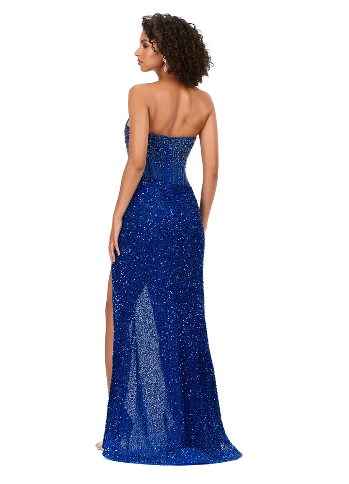 A woman models the Ashley Lauren 11238 by Ashley Lauren, a strapless floor-length royal blue sequined gown with crystal beadwork and a high slit, standing turned away to highlight its detailed back and side slit.