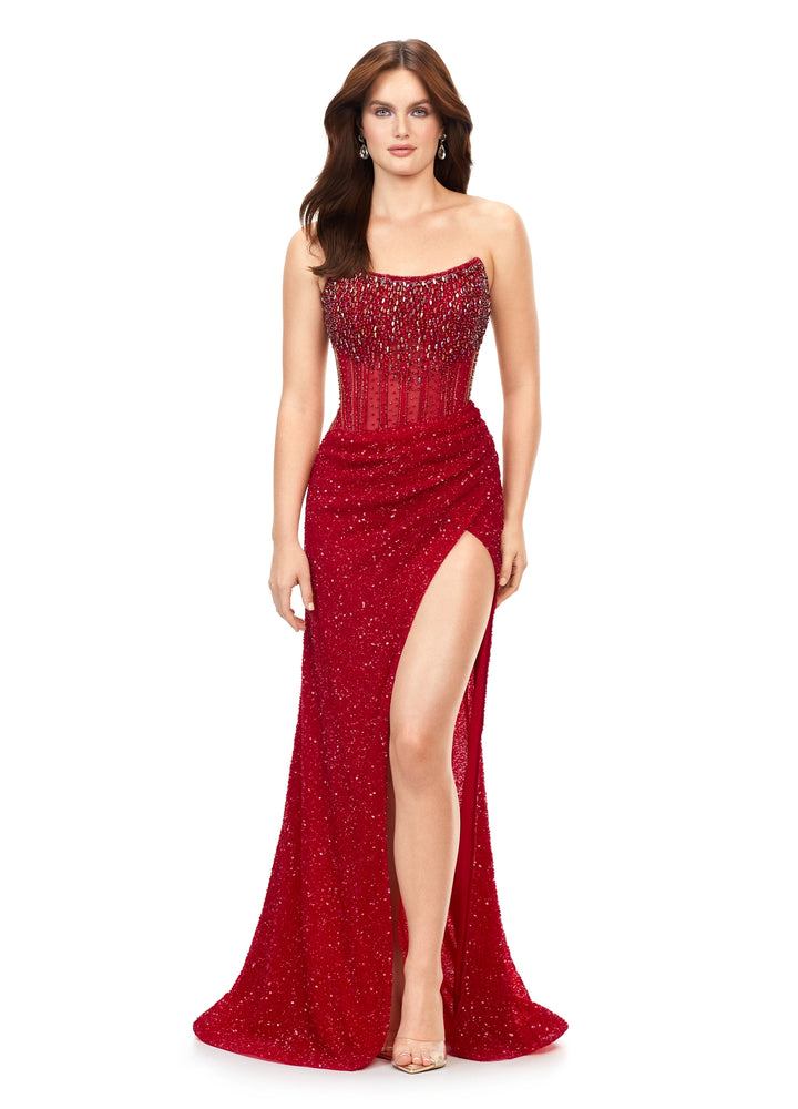 A woman wears the Ashley Lauren 11238 by Ashley Lauren, a strapless gown with crystal beadwork. The fitted bodice transitions into a long wrap skirt with a high slit. She pairs it with nude heels and has long brown hair.