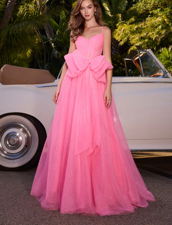 A woman wears the Ashley Lauren 11841, a sparkling bright pink ball gown with a glitter tulle skirt and large waist bow by Ashley Lauren, standing before a classic white convertible amid lush green foliage.