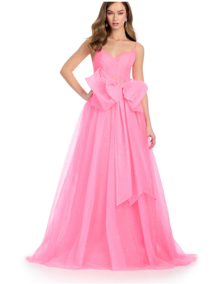 A woman models the Ashley Lauren 11841 by Ashley Lauren, a floor-length bright pink ball gown with a sweetheart neckline, thin straps, a dramatic waist bow, and a glitter tulle overlay as she poses against a white background.