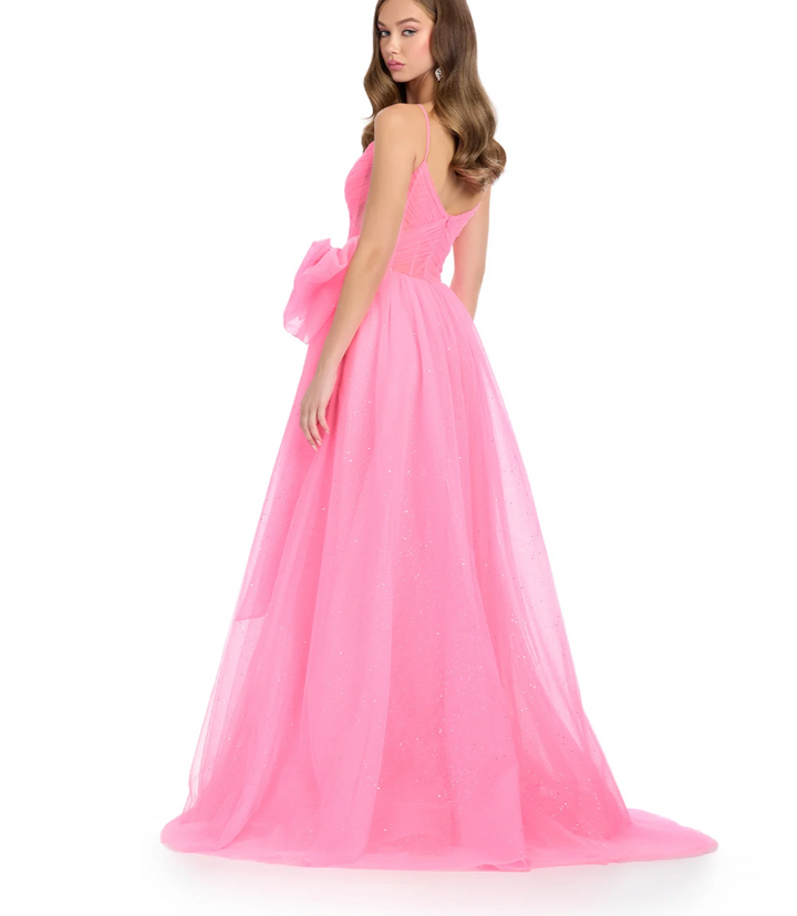 A woman models the Ashley Lauren 11841 gown by Ashley Lauren, featuring a long bright pink glitter tulle design with spaghetti straps, fitted bodice, and a large bow at the waist. She looks over her shoulder against a plain white background.