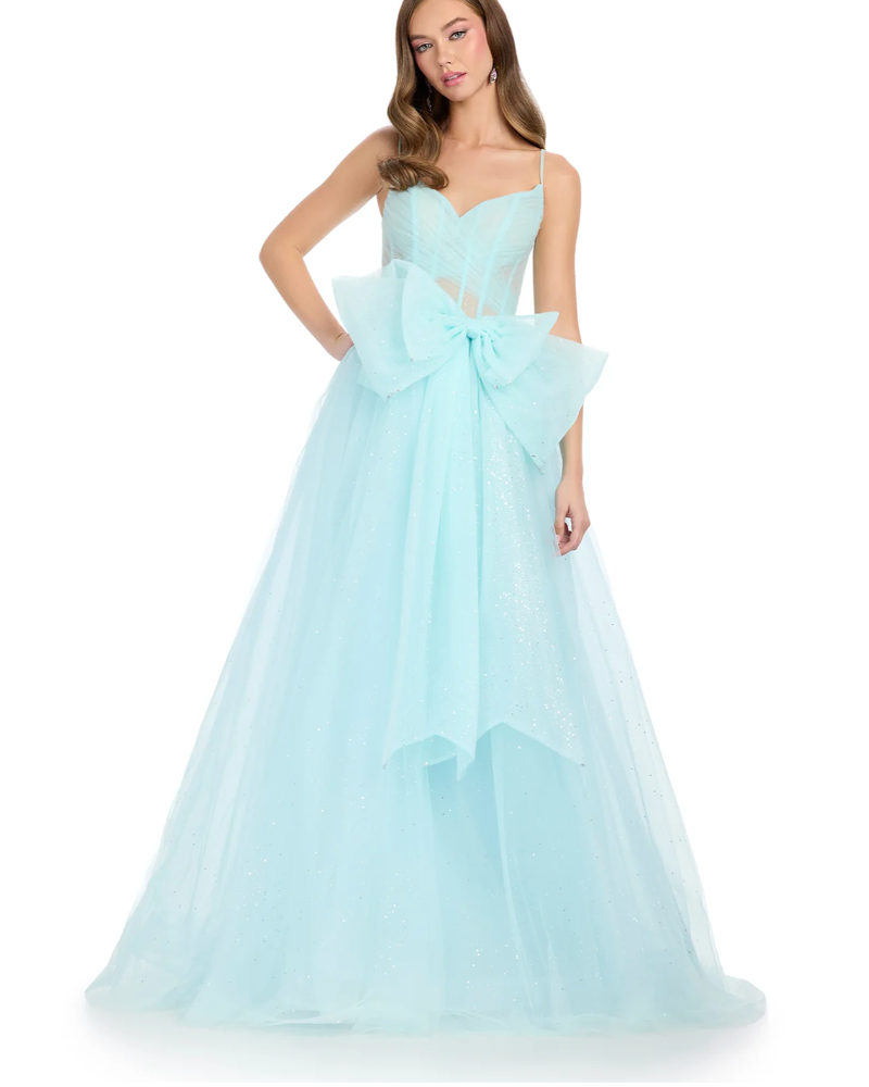A woman wears the Ashley Lauren 11841 ball gown—a light blue, floor-length dress with a sweetheart neckline, sheer bodice, spaghetti straps, waist bow accent, and glitter tulle skirt. She stands with one hand on her hip.