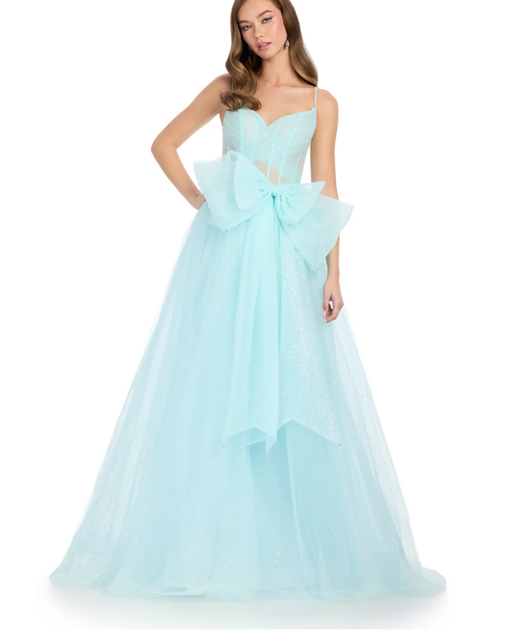 A woman wears the Ashley Lauren 11841 ball gown—a light blue, floor-length dress with a sweetheart neckline, sheer bodice, spaghetti straps, waist bow accent, and glitter tulle skirt. She stands with one hand on her hip.