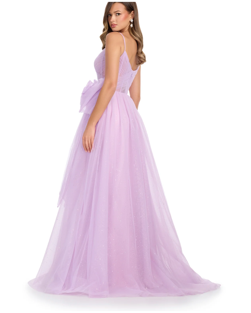 A woman in the Ashley Lauren 11841 by Ashley Lauren—a long, sleeveless light purple gown with a fitted bodice, glitter tulle skirt, and large waist bow—stands facing away while looking over her shoulder.