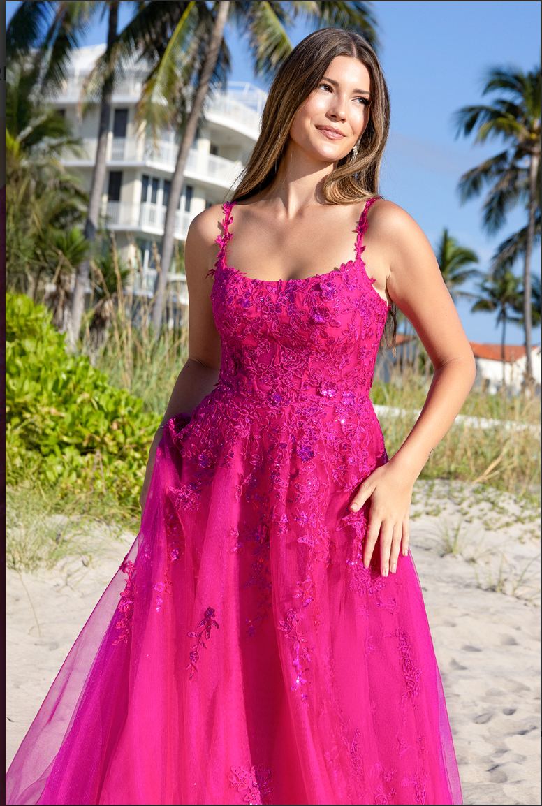 A woman wearing the Twirl Prom 4044 gown by Twirl Prom stands on a sandy beach with palm trees and a white building in the background, smiling and gazing into the distance.