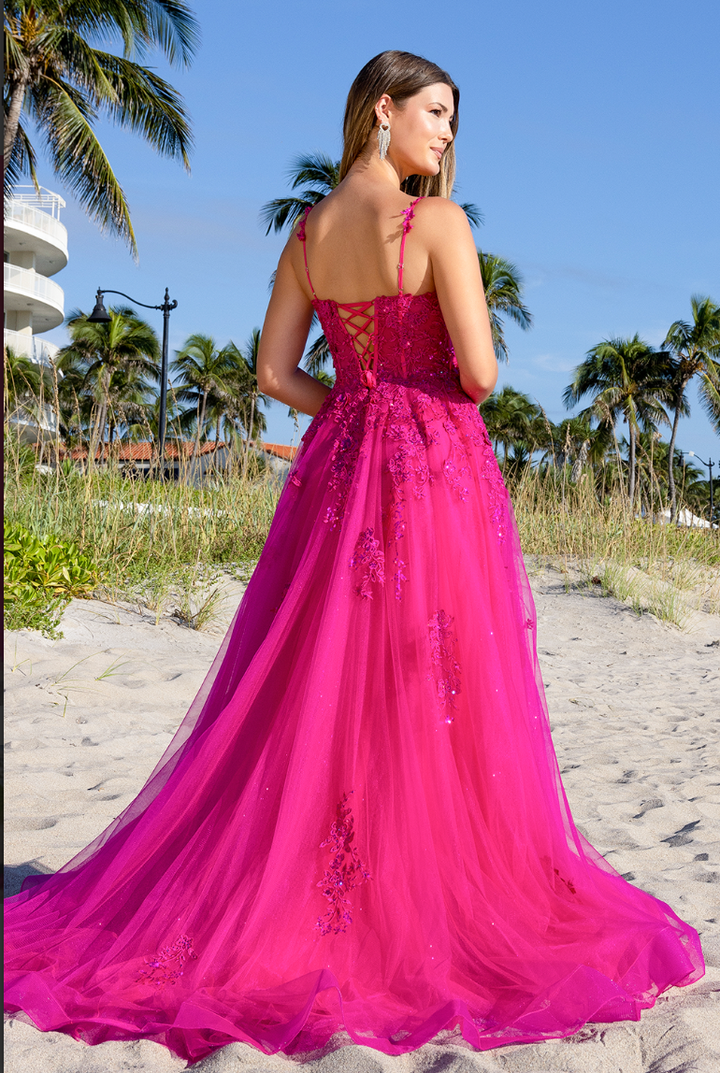 A woman stands on a sandy beach, facing away, wearing the vibrant pink Twirl Prom 4044 gown by Twirl Prom, featuring lace-up detailing and floral embellishments. Palm trees and a white building can be seen in the background.