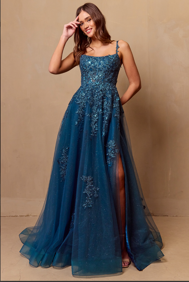 A woman in the Twirl Prom 4044 by Twirl Prom—a sparkling teal-blue, floor-length gown with floral embroidery, thin straps, and a thigh-high slit—smiles as she stands against a plain beige wall, touching her hair.