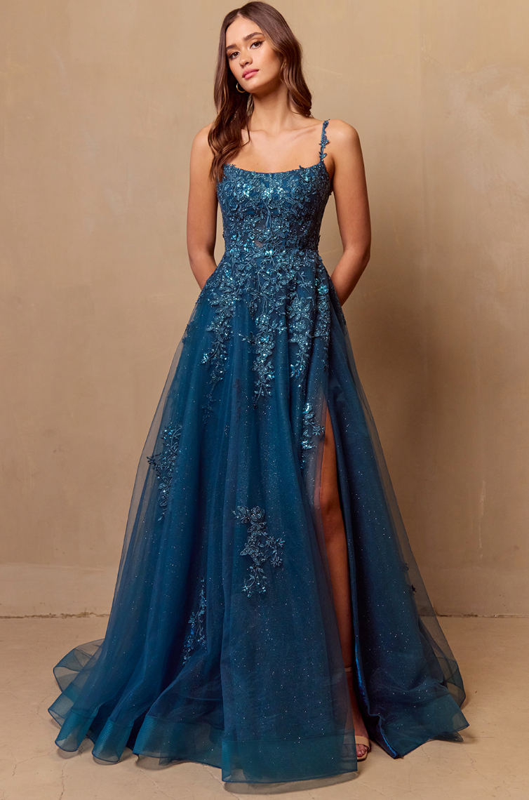 A woman poses against a beige wall in the Twirl Prom 4044 by Twirl Prom, a sleeveless teal floor-length gown with intricate beading, floral accents, and a high slit. Her hair is loose and wavy, hands behind her back.