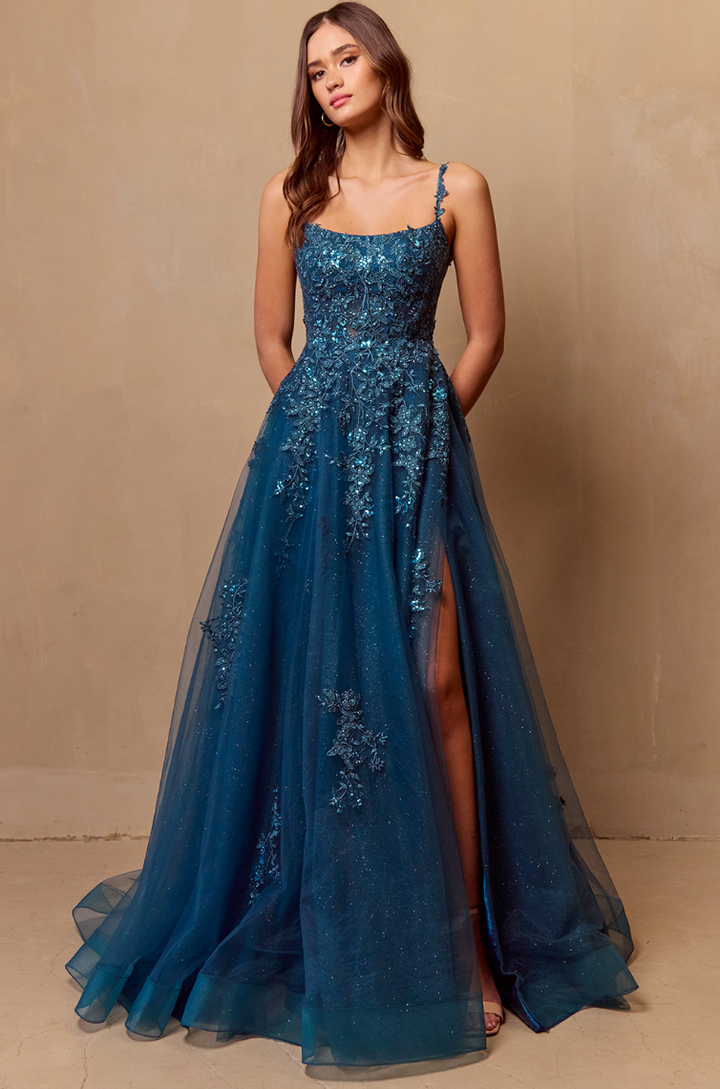 A woman poses against a beige wall in the Twirl Prom 4044 by Twirl Prom, a sleeveless teal floor-length gown with intricate beading, floral accents, and a high slit. Her hair is loose and wavy, hands behind her back.
