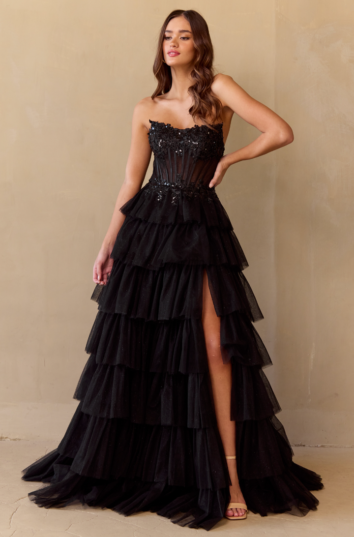 A woman models the Twirl Prom 9053 by Twirl Prom—a strapless black tiered gown with a sheer corset bodice, sequins, and a high leg slit. She stands against a beige wall with long wavy hair and open-toe heels.