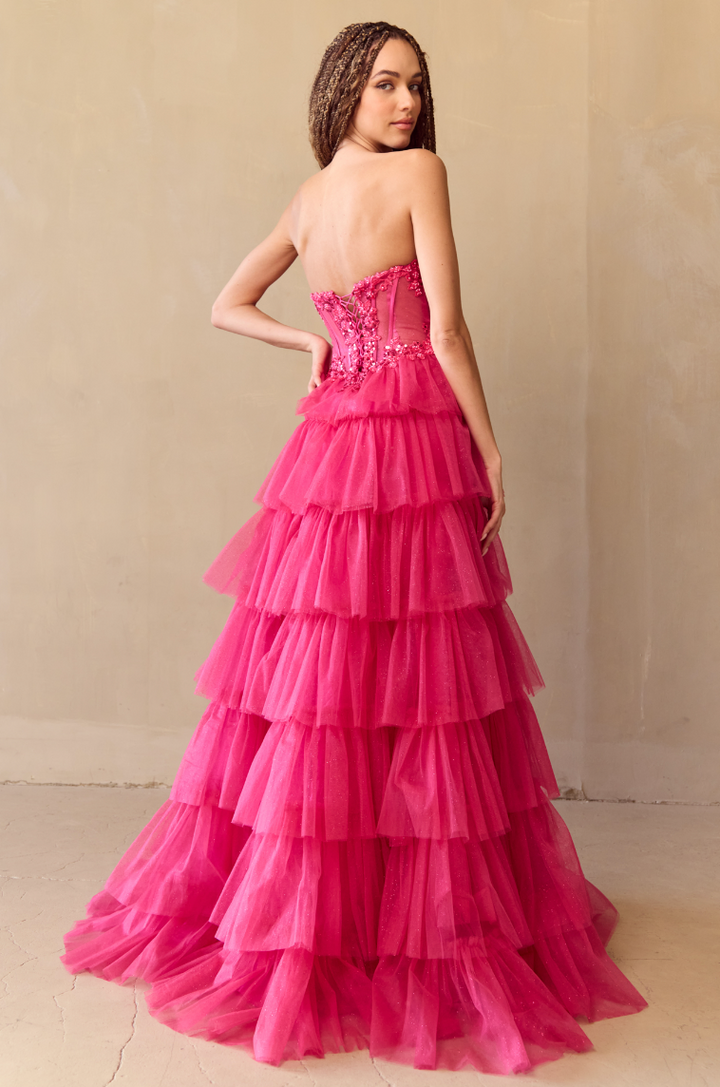 A woman with long braided hair models the Twirl Prom 9053 by Twirl Prom—a strapless, bright pink tiered tulle gown with a fitted, embellished bodice—standing against a neutral, textured wall.