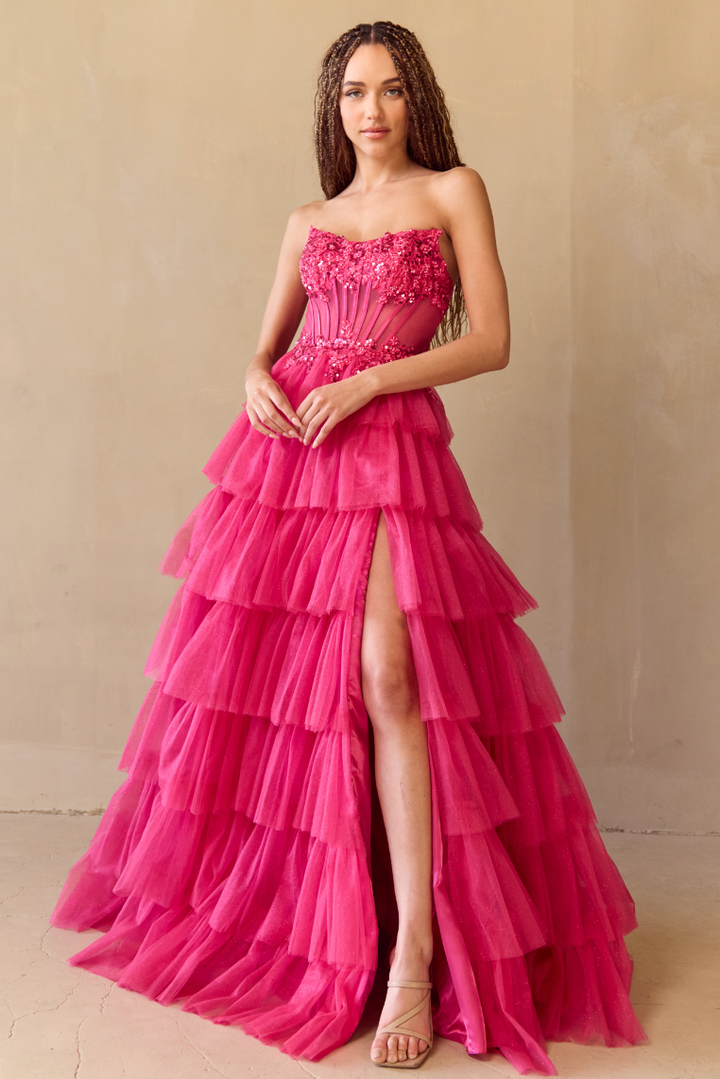 A woman models the Twirl Prom 9053 by Twirl Prom, a strapless hot pink ruffled gown with a high slit and floral bodice accents, paired with nude heels against a plain beige wall.