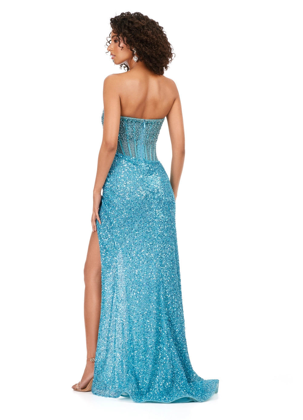 A woman models the Ashley Lauren 11238 by Ashley Lauren, featuring a strapless corset bodice with crystal beadwork, a high slit, and a floor-length wrap skirt for an elegant silhouette as she looks over her shoulder.